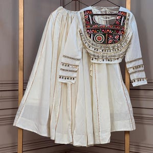 May include: A white two-piece outfit consisting of a long, flared skirt with vertical gold trim and a long-sleeved blouse. The blouse features a vibrant, multi-colored embroidered yoke with mirror work and dangling metallic accents on the sleeves.