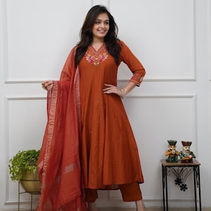 May include: A rust-colored Indian salwar kameez set. The ensemble includes a long-sleeved tunic with floral embroidery, matching pants, and a sheer scarf. The outfit is worn by a person in a well-lit room.