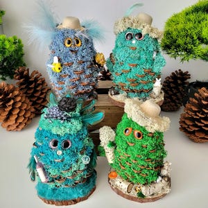 Puede incluir: Cuatro figuras caprichosas de piñas hechas a mano, cada una adornada con musgo, plumas y sombreros en miniatura. Las figuras tienen ojos saltones y están decoradas con pequeños accesorios. La paleta de colores incluye tonos de azul, verde y marrón.