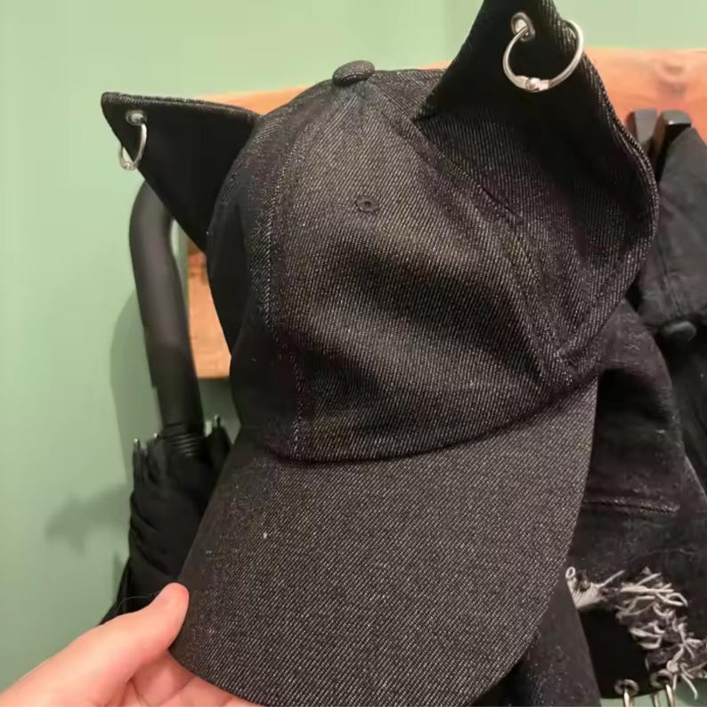 Puede incluir: Gorra de b&eacute;isbol de mezclilla negra con orejas de gato y aros plateados. La gorra tiene una visera curva y un bot&oacute;n en la parte superior. La gorra est&aacute; colgada en un estante de madera.