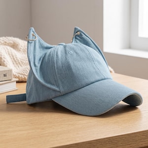 Puede incluir: Gorra de b&eacute;isbol de mezclilla azul claro con dise&ntilde;o de orejas de gato. La gorra presenta una visera curva, una correa ajustable y anillos met&aacute;licos dorados en las puntas de las orejas. El sombrero est&aacute; hecho de una tela suave y texturizada.