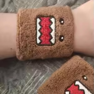 May include: Brown wristbands with an embroidered cartoon face design. The design features a red mouth, white teeth, and black eyes. The wristbands are made of a soft, absorbent material.
