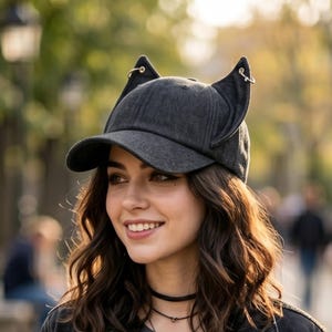 Puede incluir: Gorra de b&eacute;isbol gris oscuro con orejas de gato y peque&ntilde;os aros dorados. La gorra tiene una visera curva y est&aacute; hecha de una tela texturizada. Las orejas de gato est&aacute;n unidas a la parte superior de la gorra y se alzan.