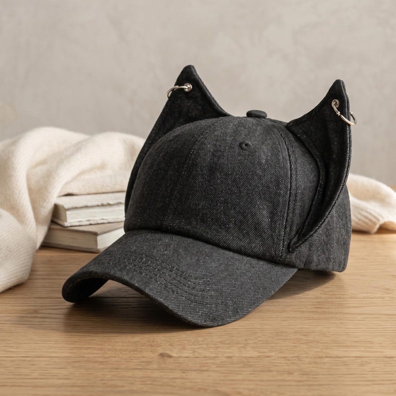 Puede incluir: Gorra de b&eacute;isbol de mezclilla gris oscuro con detalles de orejas de gato. La gorra tiene una visera curva y un bot&oacute;n en la parte superior. Aros plateados est&aacute;n unidos a las puntas de las orejas de gato. La gorra est&aacute; sobre una superficie de madera.