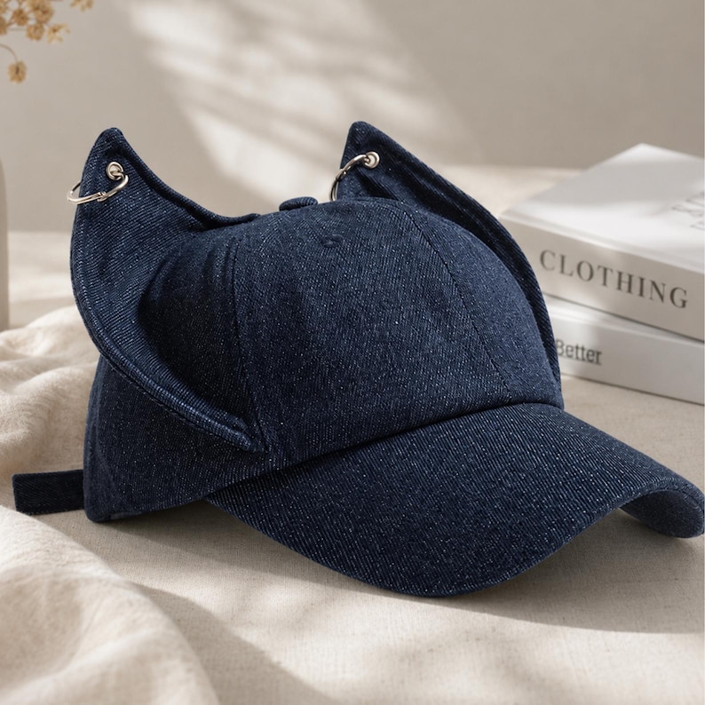 Puede incluir: Gorra de b&eacute;isbol de mezclilla azul oscuro con orejas de gato. La gorra tiene una visera curva y una correa de tela ajustable en la parte trasera. Las orejas de gato est&aacute;n sujetas con anillos de metal plateado. La palabra "CLOTHING" es visible en un libro en el fondo.