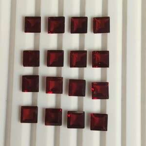 May include: Sixteen square-cut, deep red gemstones arranged on a white surface. The gemstones are evenly spaced and reflect light, showcasing their faceted surfaces. The gems appear to be of similar size and shape.