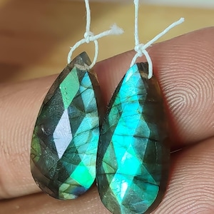 Puede incluir: Dos piedras preciosas de labradorita facetadas en forma de lágrima. Las piedras son una mezcla de gris oscuro, verde y azul, con destellos de color iridiscente. Cada piedra está ensartada en un cordón blanco.