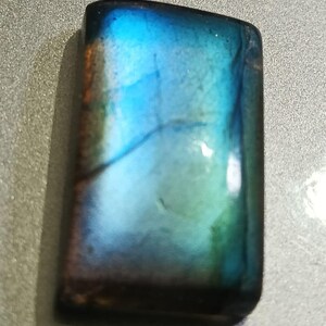 May include: A rectangular, polished labradorite gemstone with a captivating play of color. The stone displays iridescent hues of blue, green, and hints of gold, creating a shimmering effect. The edges are smooth and the overall shape is rectangular.