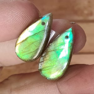 Puede incluir: Dos piedras preciosas de labradorita en forma de lágrima. Las piedras muestran una iridiscencia verde y azul vibrante, con un punto oscuro cerca de la parte superior. Las superficies pulidas reflejan la luz, mostrando patrones naturales. Ideal para joyería.