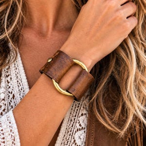 May include: A wide cuff bracelet featuring two pieces of distressed brown leather connected by a polished gold-tone metal ring. The leather sections are secured with a small metal stud, creating a structured design that wraps around the wrist.