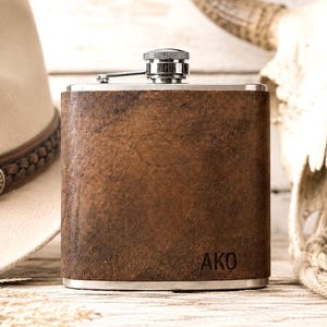 May include: A rectangular stainless steel flask wrapped in a textured brown leather-like material. The flask features a silver-colored metal screw-top cap and the letters AKO embossed in the bottom right corner of the front surface.