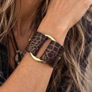 Könnte beinhalten: Ein breites Armband mit einem braun-schwarzen Tierfellmuster auf strukturiertem Material. Das Armband wird durch einen polierten goldfarbenen Metallring zusammengehalten, der zwei separate Abschnitte des Musters mit kleinen goldenen Nieten verbindet.