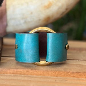 K&ouml;nnte beinhalten: Ein breites Armband mit einem strukturierten, t&uuml;rkisfarbenen Lederband, das mit zwei messingfarbenen Metallnieten befestigt ist. Das Lederband ist durch einen gro&szlig;en, polierten, goldfarbenen kreisf&ouml;rmigen Metallring gezogen, der das Zentrum bildet.