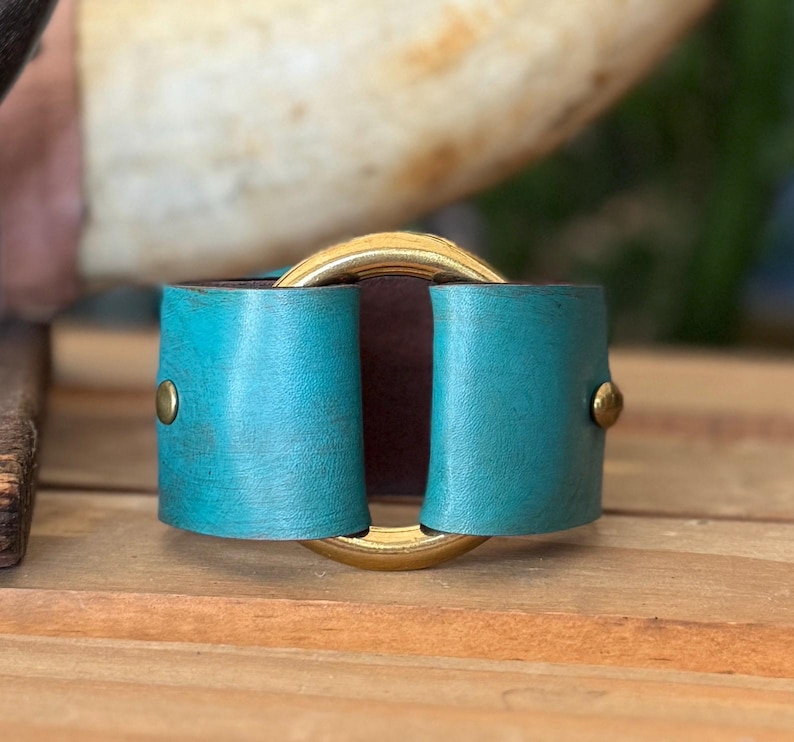 K&ouml;nnte beinhalten: Ein breites Armband mit einem strukturierten, t&uuml;rkisfarbenen Lederband, das mit zwei messingfarbenen Metallnieten befestigt ist. Das Lederband ist durch einen gro&szlig;en, polierten, goldfarbenen kreisf&ouml;rmigen Metallring gezogen, der das Zentrum bildet.