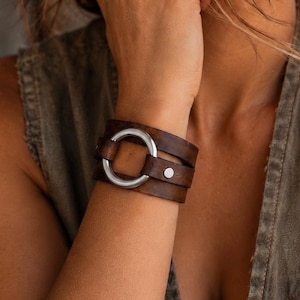 Könnte beinhalten: Ein breites Armband aus dunkelbraunem Leder mit einem glatten, silberfarbenen Metallring in der Mitte. Das Lederband umschließt das Handgelenk und wird an der Seite mit einem kleinen, silberfarbenen Metallnietknopf sicher verschlossen.