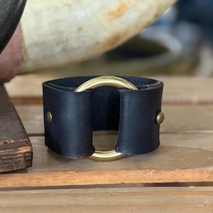K&ouml;nnte beinhalten: Ein schwarzes Lederarmband mit einem glatten, polierten goldfarbenen Metallring in der Mitte. Das Lederband ist auf beiden Seiten des Rings mit kleinen, runden, goldfarbenen Metallnieten befestigt, was ein strukturiertes, kreisf&ouml;rmiges Design ergibt.