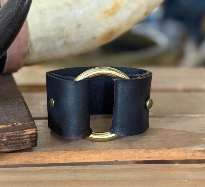 K&ouml;nnte beinhalten: Ein schwarzes Lederarmband mit einem glatten, polierten goldfarbenen Metallring in der Mitte. Das Lederband ist auf beiden Seiten des Rings mit kleinen, runden, goldfarbenen Metallnieten befestigt, was ein strukturiertes, kreisf&ouml;rmiges Design ergibt.