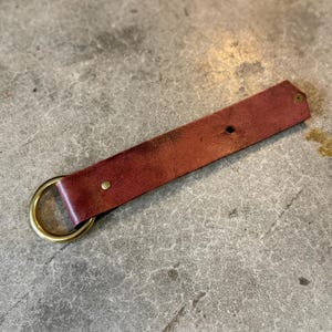 K&ouml;nnte beinhalten: Ein rechteckiger Streifen aus rotbraunem Leder, der um einen runden Messingring gefaltet ist. Der Lederriemen ist mit zwei kleinen Messingnieten befestigt, eine in der N&auml;he des Rings und eine am spitzen Ende, mit sichtbarer nat&uuml;rlicher Maserung.
