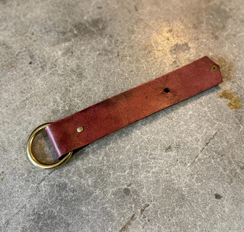 K&ouml;nnte beinhalten: Ein rechteckiger Streifen aus rotbraunem Leder, der um einen runden Messingring gefaltet ist. Der Lederriemen ist mit zwei kleinen Messingnieten befestigt, eine in der N&auml;he des Rings und eine am spitzen Ende, mit sichtbarer nat&uuml;rlicher Maserung.