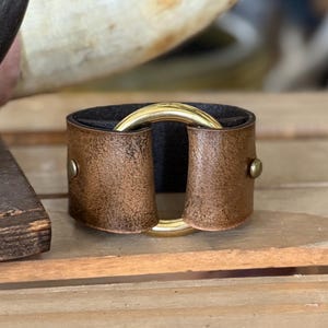 K&ouml;nnte beinhalten: Ein breites Armband aus abgenutztem braunem Leder mit dunkler Innenseite. Das Leder ist an den Seiten mit zwei kleinen messingfarbenen Metallnieten befestigt und hat einen gro&szlig;en, polierten goldfarbenen Metallring in der Mitte.