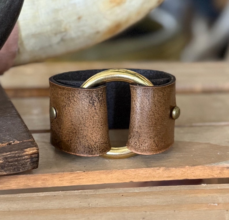 K&ouml;nnte beinhalten: Ein breites Armband aus abgenutztem braunem Leder mit dunkler Innenseite. Das Leder ist an den Seiten mit zwei kleinen messingfarbenen Metallnieten befestigt und hat einen gro&szlig;en, polierten goldfarbenen Metallring in der Mitte.