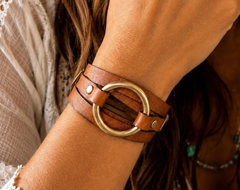 May include: A brown leather cuff bracelet featuring multiple layered leather straps secured by small metal rivets. A large, circular, antique-toned brass ring serves as the central focal point, connecting the leather bands together in a wrap-around design.