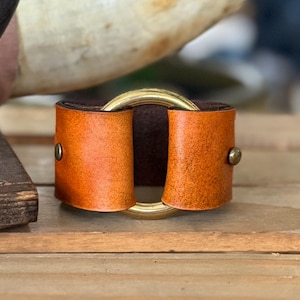 K&ouml;nnte beinhalten: Ein breites Lederarmband mit einer leuchtend orangefarbenen Au&szlig;enseite und einer dunkelbraunen Innenseite. Das Leder wird durch einen zentralen Ring aus poliertem goldfarbenem Metall und zwei kleine messingfarbene Nieten gehalten.