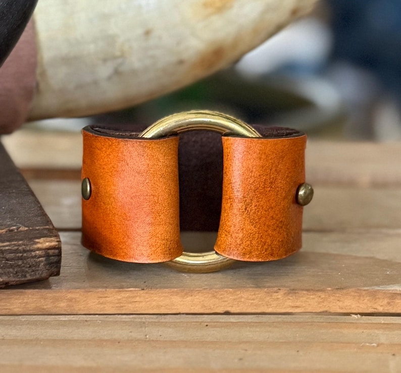 K&ouml;nnte beinhalten: Ein breites Lederarmband mit einer leuchtend orangefarbenen Au&szlig;enseite und einer dunkelbraunen Innenseite. Das Leder wird durch einen zentralen Ring aus poliertem goldfarbenem Metall und zwei kleine messingfarbene Nieten gehalten.