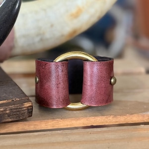 K&ouml;nnte beinhalten: Ein breites Armband aus dunkelrotem Leder mit Used-Optik, das in der Mitte einen gro&szlig;en, polierten goldfarbenen Metallring aufweist. Die Lederteile sind auf beiden Seiten mit kleinen, runden, goldfarbenen Metallnieten am Ring befestigt.