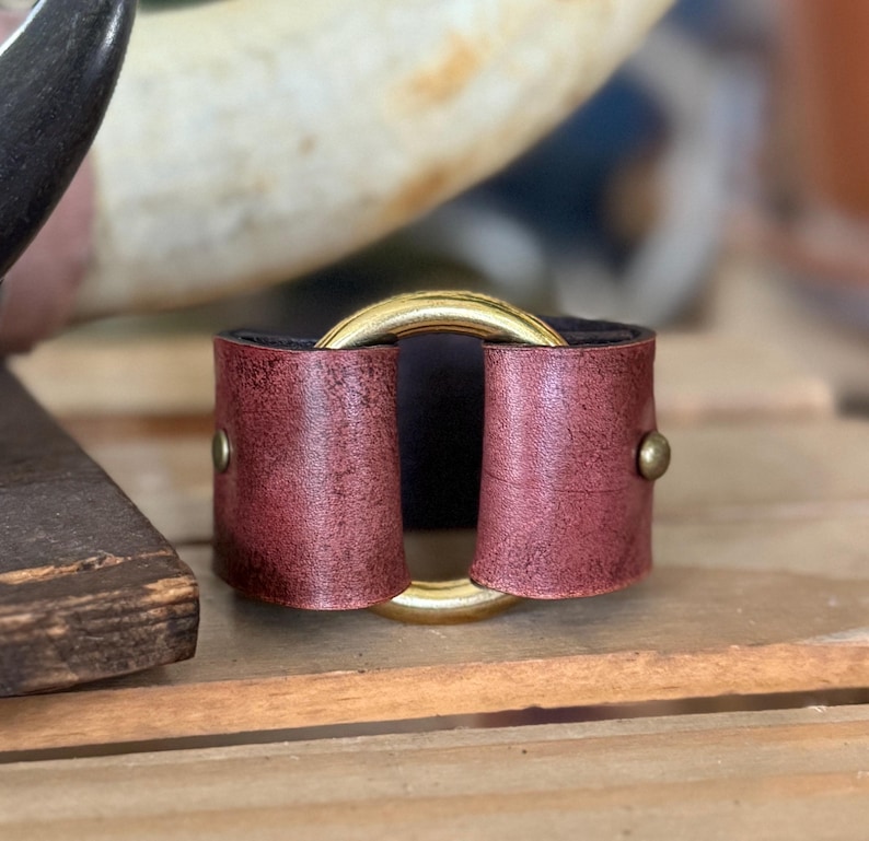 K&ouml;nnte beinhalten: Ein breites Armband aus dunkelrotem Leder mit Used-Optik, das in der Mitte einen gro&szlig;en, polierten goldfarbenen Metallring aufweist. Die Lederteile sind auf beiden Seiten mit kleinen, runden, goldfarbenen Metallnieten am Ring befestigt.