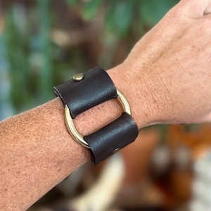 K&ouml;nnte beinhalten: Ein dunkelbraunes Lederarmband mit einem glatten, polierten goldfarbenen Metallring in der Mitte. Das Lederband ist auf beiden Seiten des zentralen Rings mit kleinen, runden messingfarbenen Metallnieten befestigt, was ein strukturiertes Design ergibt.