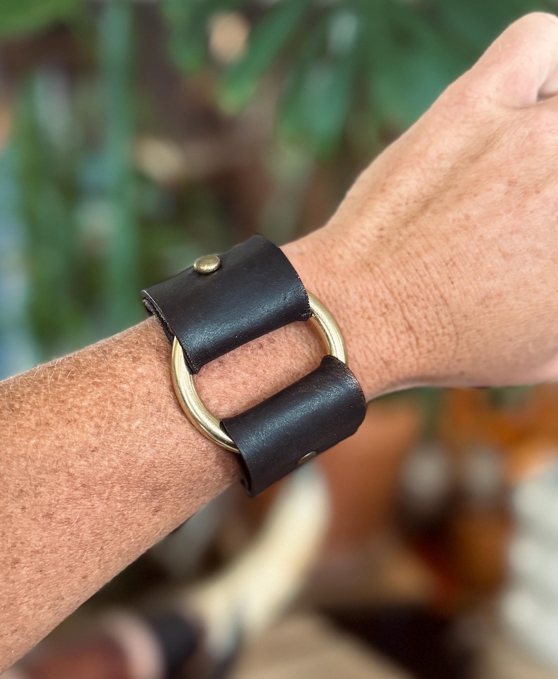K&ouml;nnte beinhalten: Ein dunkelbraunes Lederarmband mit einem glatten, polierten goldfarbenen Metallring in der Mitte. Das Lederband ist auf beiden Seiten des zentralen Rings mit kleinen, runden messingfarbenen Metallnieten befestigt, was ein strukturiertes Design ergibt.