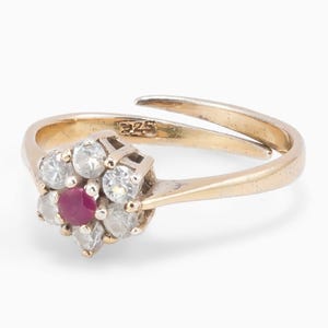 Op de afbeelding: Een goudkleurige ring met open band en een bloemvormige zetting. Het middenstuk is een ronde, dieproze edelsteen omringd door zes heldere, gefacetteerde stenen. De binnenkant van de ring is gestempeld met 925, wat duidt op sterling zilver.