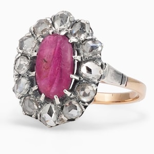 May include: A ring featuring a central oval cabochon-cut deep pink ruby surrounded by a halo of twelve rose-cut diamonds. The setting is crafted from silver and gold, with a polished gold band that tapers toward the back of the ring.