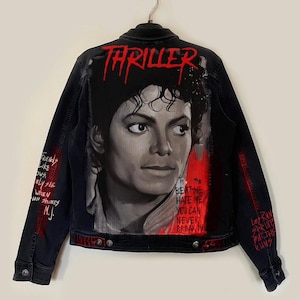 Può includere: Giacca di jeans nera con un ritratto di Michael Jackson e la parola "THRILLER" in rosso. La giacca presenta dettagli rossi e testo scritto a mano sulle maniche, tra cui "Beat Me, Hate Me, You Can Never Break Me."