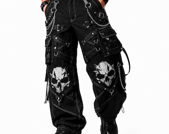May include: Black cargo pants featuring a white skull graphic on each leg, multiple utility pockets, silver-toned metal chains, D-rings, and adjustable straps. The pants include a black belt with silver studs and are paired with black platform boots.