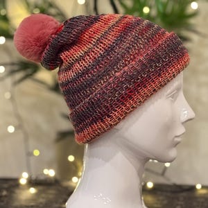 Könnte beinhalten: Eine gestrickte Beanie-Mütze mit einem rosa Bommel. Die Mütze hat ein gestreiftes Muster in Rot-, Orange- und Dunkelgrautönen. Die Mütze wird auf einem weißen Schaufensterkopf präsentiert, der ihr Design und ihre Textur zeigt.