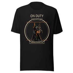 May include: A black short-sleeved t-shirt featuring a graphic of a Doberman dog head inside a circular frame. The text ON DUTY BY INSTINCT appears above the circle, and DOBERMAN WORKING CLUB is printed in a rectangular box below the circle.