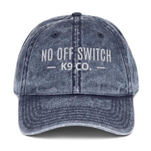 May include: A navy blue acid-wash baseball cap featuring the embroidered white text NO OFF SWITCH K9 CO. centered on the front panel. The cap has a curved brim, a button on the top crown, and stitched eyelets for ventilation on each panel.