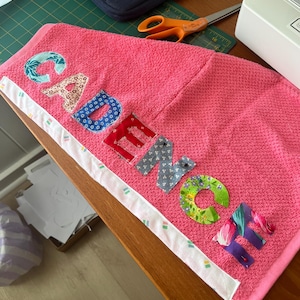 Peut inclure: Une serviette en tissu éponge rose vif portant le nom CADENCE appliqué en lettres de tissus à motifs variés. Chaque lettre est cousue sur la serviette, certaines étant fixées par des épingles. Un liseré blanc à motifs verts et orange borde un côté.