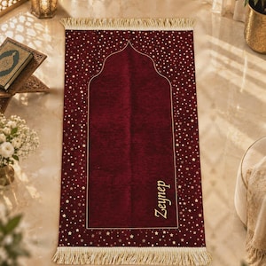 May include: A rectangular prayer rug in deep burgundy velvet featuring a gold arched mihrab outline and a scattered gold star pattern. The rug includes cream-colored fringe on the top and bottom edges and the name Zeynep embroidered in gold.