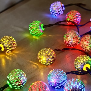 May include: A string of decorative globe lights, each sphere patterned and emitting a warm glow. The lights display a variety of colours, including green, yellow, red, and purple. The lights are strung on a dark wire, ideal for festive decoration.