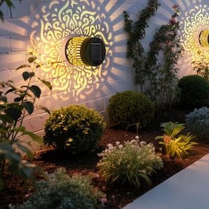 Pode incluir: Luzes de parede externas movidas a energia solar com intrincados padrões recortados. As luzes projetam um padrão decorativo na parede de tijolos brancos. As luzes são cilíndricas com um painel solar preto. O jardim apresenta arbustos e plantas verdes.