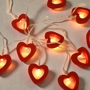 Könnte beinhalten: Eine Lichterkette mit roten herzförmigen Lichtern, die ein warmes, gelbes Leuchten ausstrahlen. Die Lichter sind an einem dünnen, transparenten Draht befestigt. Ideal für eine romantische Atmosphäre, z.B. zum Valentinstag.