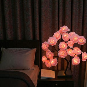 May include: A decorative rose tree lamp with multiple illuminated pink rose blossoms. The lamp is on a black base and sits on a nightstand next to a bed with a pillow and a book.