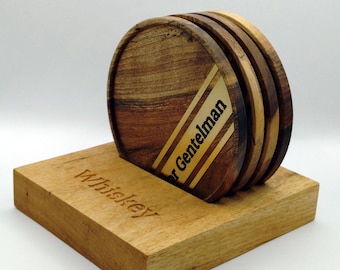 May include: A set of four wooden coasters with a holder. The coasters are round with a striped design and the text "r Gentelman". The holder is rectangular with the word "Whiskey" engraved on it.