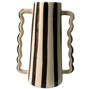 Puede incluir: Un jarrón alto de cerámica con base crema y rayas verticales negras. Presenta dos asas onduladas. El diseño es sencillo y moderno, adecuado para varios estilos de decoración de interiores. Altura aproximada de 30 cm.