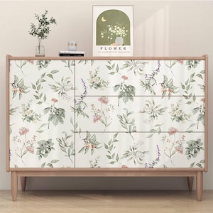 Op de afbeelding: Een houten dressoir met een bloemenpatroon op de lades. Het patroon bevat verschillende bloemen en bladeren in roze, groene en witte tinten. Het dressoir heeft een lichtbruin houten frame en poten. Een ingelijste afbeelding met het woord "FLOWER" staat erop.