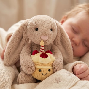 May include: A plush, light brown bunny toy holding a cupcake with a lit candle. The cupcake has red cherry-like accents and a smiling face. The bunny is resting on a cream-colored blanket.