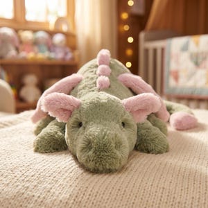Pode incluir: Um brinquedo de pel&uacute;cia verde em forma de dinossauro com detalhes rosa nas orelhas, espinhos e p&eacute;s. O brinquedo macio est&aacute; deitado em um cobertor de malha creme. O dinossauro tem uma express&atilde;o amig&aacute;vel e &eacute; projetado para crian&ccedil;as.